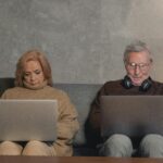 An elderly couple sitting on a sofa at home, each using a laptop. Modern technology engagement.